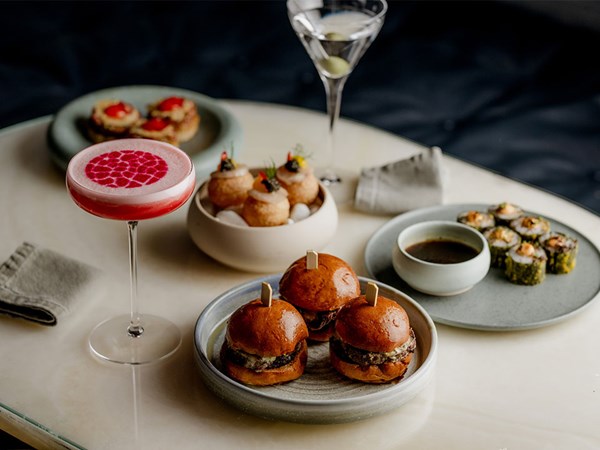 Three beef burger sliders on a plate, a pink cocktail in a glass and three sushi rolls served on a plate